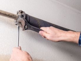 A person uses a wrench and a screwdriver to adjust or repair a metal garage door torsion spring mounted on the ceiling.