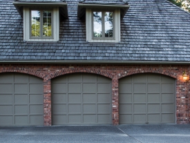 A brick house with three closed gray garage doors and two upper windows with a shingled roof. A single wall light is on near the right side of the garage.