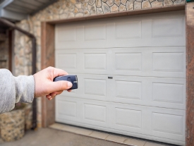 A person’s hand holds a remote control in front of a closed white garage door, appearing to open or close it. The garage is part of a stone house exterior.