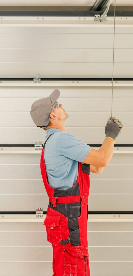 A technician in red overalls and a gray cap adjusts a cable on a closed garage door, looking up while working.